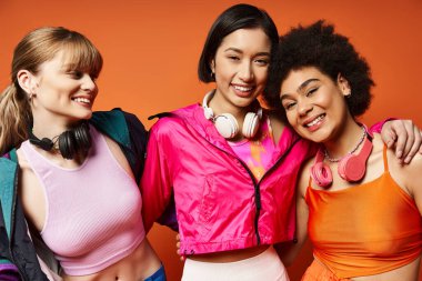 A diverse group of women, including Caucasian, Asian, and African American, standing close together against an orange studio background. clipart