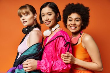 Farklı etnik kökenlere ve geçmişlere sahip üç kadın bir stüdyoda turuncu bir zemine karşı yan yana duruyorlar..