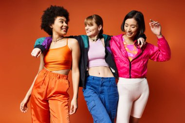 Farklı etnik kökenlere sahip üç kadın bir stüdyo ortamında birbirine yakın duruyor, canlı turuncu arkaplan arasında çeşitlilik sergiliyorlar..