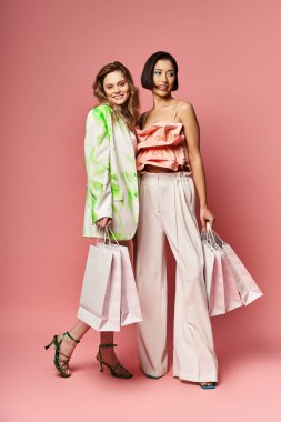 Two women, one Caucasian, one African American, stand with shopping bags against a pink studio background. clipart