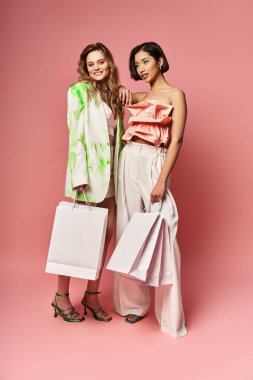 İki çok kültürlü kadın, canlı pembe bir arka planda alışveriş torbalarıyla yan yana duruyorlar..