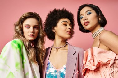 Farklı etnik kökenlere sahip üç kadın pembe bir arkaplan önünde birlikte duruyor, güzellik ve çeşitlilik sergiliyorlar..