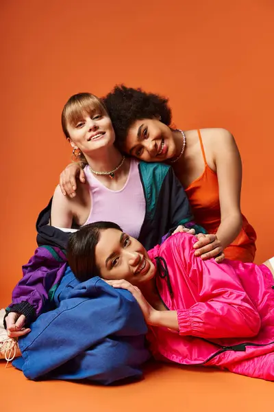 A group of multicultural women, including Caucasian, Asian, and African American, joyfully laying on top of each other on an orange background.