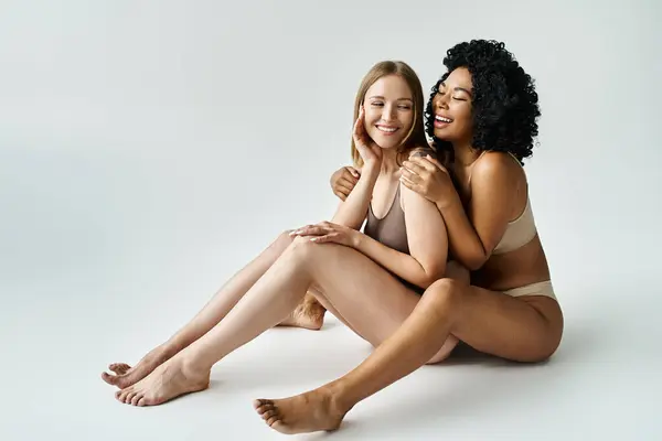 stock image Two diverse women in cozy pastel underwear seated together against a white background.