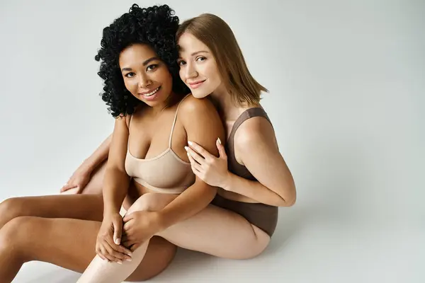 stock image Two diverse women in cozy pastel underwear sitting together on a white background.