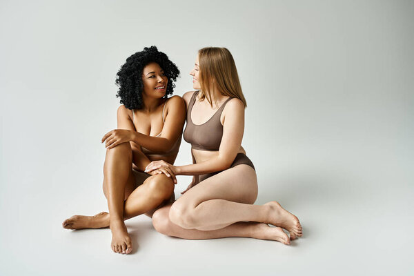Two diverse women in cozy pastel underwear, sitting closely next to each other on a white background.