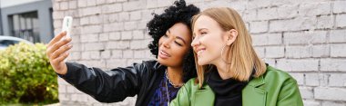 Two attractive diverse women in casual attire taking a selfie with a cell phone. clipart