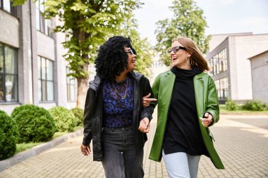 Two stylish women walking and chatting animatedly along a city sidewalk. clipart