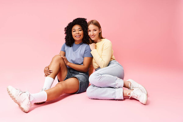Two diverse women in casual attire sitting on the ground, posing for a picture.