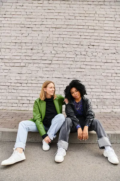 stock image Two diverse women in casual attire sit on curb by brick wall.