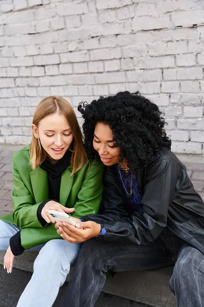 stock image Two diverse women in casual attire sitting on the ground, engrossed in a cell phone.