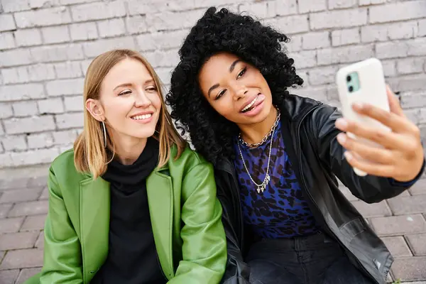 stock image Two women in casual attire sitting on the ground, capturing a moment with a selfie.