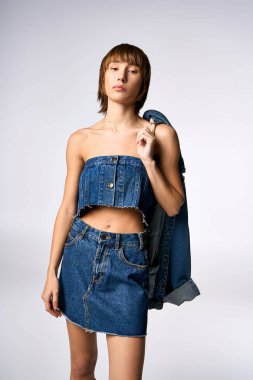 Kısa saçlı genç bir kadın, stüdyo ortamında kot elbise ve ceketle güven içinde poz veriyor..