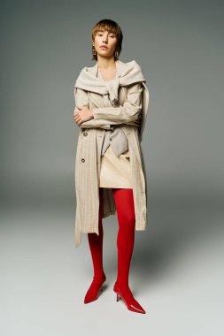 A young woman with short hair is strikingly dressed in a trench coat and vibrant red tights in a studio setting. clipart