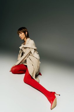 A stylish young woman with short hair poses boldly in a trench coat and vibrant red tights in a studio setting. clipart