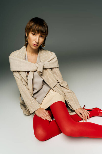 A young woman with short hair sits on the floor in a studio setting, donning red tights with elegance and poise.