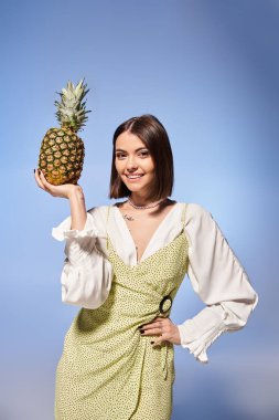 Esmer saçlı genç bir kadın stüdyo ortamında ananası yüzüne doğru tutar..