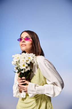 Kumral saçlı, güneş gözlüğü takan genç bir kadın, sakin bir stüdyoda elinde papatya buketi tutuyor..