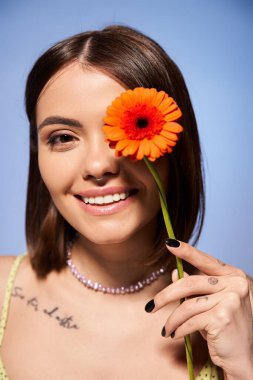 A brunette woman delicately holds a flower in front of her face in a studio setting, connecting with nature. clipart