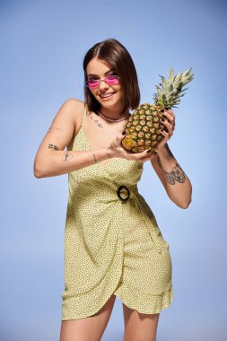 Sarı elbiseli esmer bir kadın stüdyo ortamında ananas tutuyor..