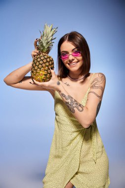 Esmer saçlı genç bir kadın stüdyoda poz veriyor. Parlak sarı bir elbise giyiyor, elinde canlı bir ananas tutuyor..