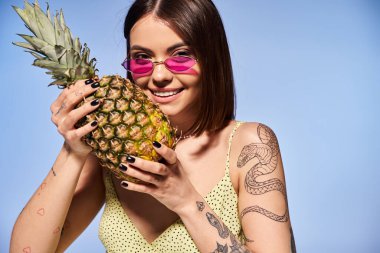 Stüdyoda son moda güneş gözlüğü takan esmer saçlı şık bir genç kadın ananas tutuyor..