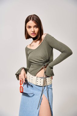 Esmer kadın kendinden emin bir şekilde elleri etekli, akıcı kalçaları üzerinde duruyor. Stüdyo ortamında zarafet ve duruş sergiliyor..