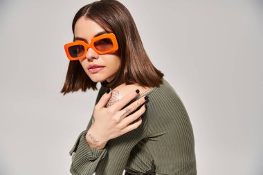 Genç bir esmer kadın canlı yeşil bir kazak ve turuncu güneş gözlüğü ile stüdyoda poz veriyor..