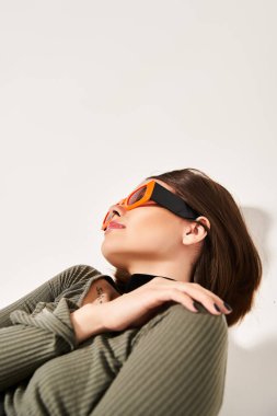 Esmer saçlı genç bir kadın, canlı yeşil bir süveter ve şık turuncu güneş gözlüğü takarak stüdyoda şık poz veriyor..