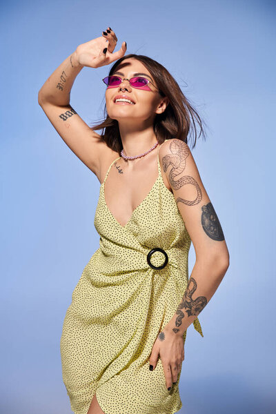A brunette woman in a stunning yellow dress proudly shows off her intricate arm tattoos in a studio setting.