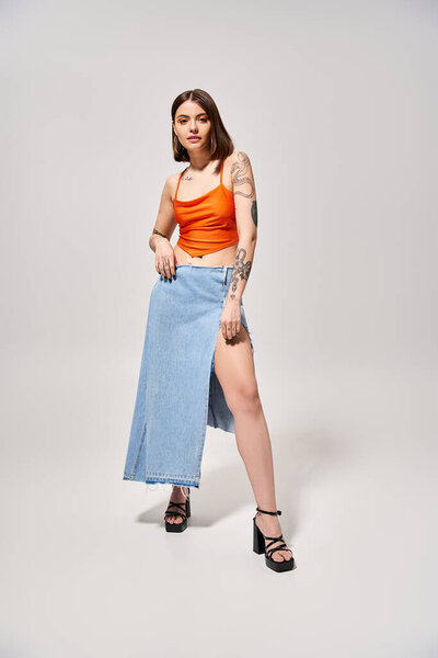 A brunette woman poses gracefully in a studio wearing an orange top and blue skirt, exuding elegance and vibrancy.