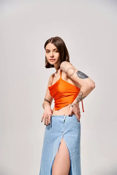 stock image A young woman with brunette hair is standing gracefully in a studio, wearing an orange top and a flowing blue skirt.