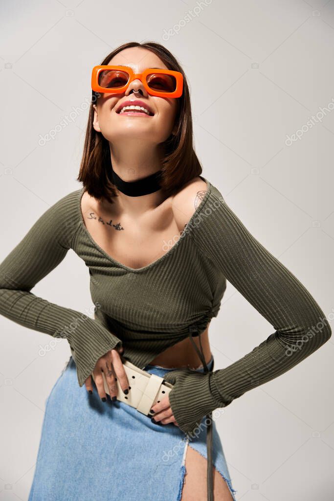 A fashionable young brunette woman confidently wears sunglasses and a stylish skirt in a studio setting.