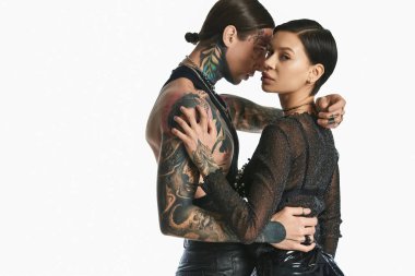Genç, dövmeli bir çift, stüdyoda gri bir arka plana karşı sıcak bir kucaklaşmayı paylaşıyor, sevgi ve yakınlık gösteriyor..