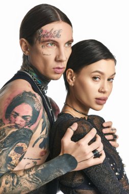 Kollarında zarif dövmeleri olan genç bir adam ve kadın gri bir arka plana karşı bir stüdyoda güven içinde poz veriyorlar..