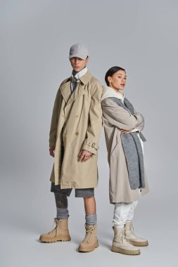 A stylish young couple standing next to each other in trench coats, posing in a studio against a grey background. clipart