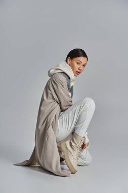 Yerde oturan genç bir stil kadın, gri bir stüdyo arka planı karşısında, bir trençkot içinde şık görünüyor.