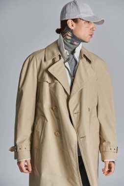 A young, tattooed man exudes mystery and intrigue in a trench coat, donning a stylish hat against a grey studio backdrop. clipart
