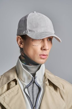 Genç, dövmeli bir adam şık bir trençkotla moda bir şapkayla birlikte gri bir stüdyo arkaplanına karşı karizma yayıyor..