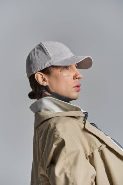 Genç, dövmeli bir adam, gri bir stüdyo arkaplanına karşı trençkot, şapka ve ceketle güvenle poz veriyor..