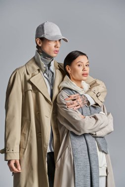 Genç bir adam ve bir kadın, gri bir stüdyo arkaplanına karşı siperler ve entelektüellik içinde yan yana durur..