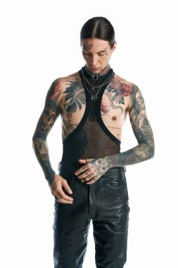 A young man with striking tattoos and piercings on his chest poses in a studio against a grey background. clipart