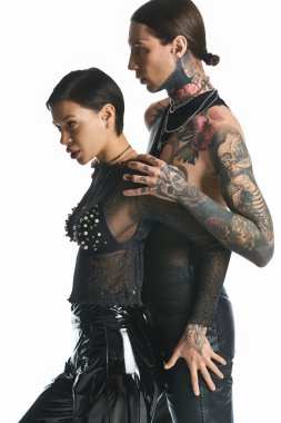 A young, stylish couple with intricate tattoos on their bodies posing in a studio against a grey background. clipart