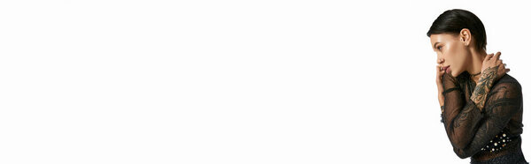 Женщина с татуировками на руке нежно касается своего лица, размышляя о глубоких эмоциях в студии на сером фоне.