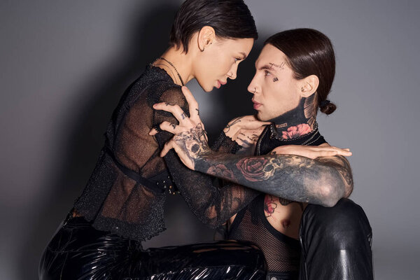 A young tattooed couple sitting together in a studio, sharing a moment of passion and intimacy against a grey background.