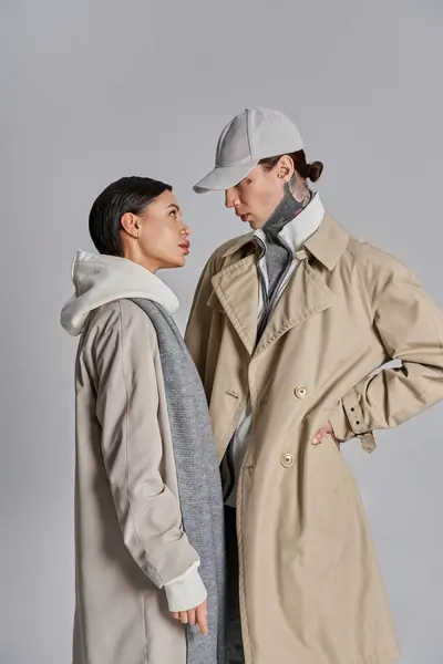 Stock image A young stylish couple stands side by side in trench coats, exuding sophistication and charm in a studio against a grey backdrop.