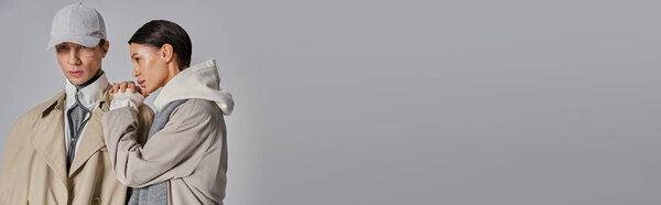 Молодой человек и женщина, стильно одетые в плащи, стоят бок о бок в студии на сером фоне.