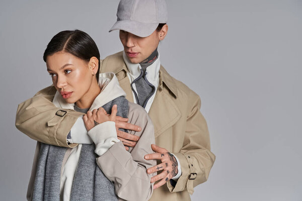 A young stylish couple, the man holding the woman tenderly in his arms, captured in a moment of intimacy on a grey background.