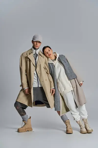 一对年轻貌美的情侣在一间灰色背景的工作室里肩并肩站在战壕外套上 — 图库照片