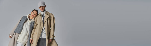 Молодой человек и женщина, одетые в стильные плащи, стоят бок о бок в студии на сером фоне.